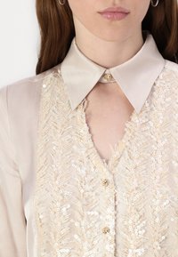 Blusa beige con colletto fitted, caratterizzata da un ricamo in paillettes intricati con motivo a felce e bottoni dorati lungo la placca.