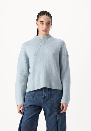 Pull en maille côtelée bleu clair avec un col haut et des épaules tombantes, associé à un pantalon cargo bleu foncé avec des poches latérales et des détails en bouton.