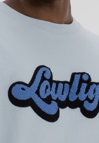 Světle modré tričko s kulatým výstřihem, na přední straně s černě a modře texturovaným výšivkovým logem "Lowlight". Měkký bavlněný materiál.
