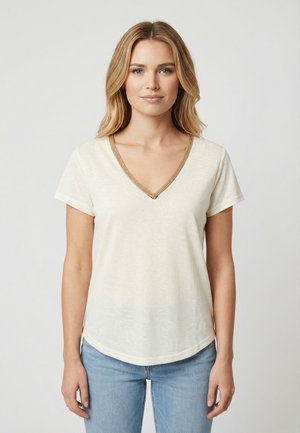 Joven mujer con cabello rubio que lleva una camiseta de cuello en V color crema con ribete marrón y jeans azul claro, de pie frente a un fondo gris liso.