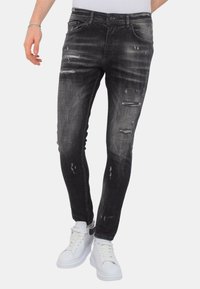Zwarte gescheurde jeans met een vervaagde afwerking, voorzien van meerdere versleten details en een slim fit. Gecombineerd met witte sneakers.