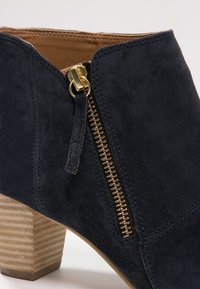 Granatowe zamszowe botki za kostkę z bocznym zamkiem, z metalowym uchwytem w złotym odcieniu i piętą wykonaną z warstwowego drewna. Gładka tekstura, wyraziste linie projektu.