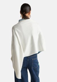 Bílý pletený šál přehozený přes ramena, s hladkým povrchem. Nosený přes světle modrou košili a v kombinaci s tmavými džíny.