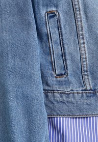 Jeansjacka i ljusblått med kontrasterande sömmar, en bröstficka och en texturerad yta, lager på lager över ett randigt blått tyg.