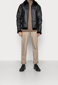 Giacca di pelle nera con collare in pelliccia sintetica, superficie testurizzata, chiusura con zip, abbinata a pantaloni beige e sneaker nere.