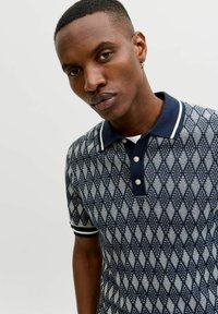 Man wearing a navy blue and white diamond-patterned polo shirt with collar and buttons, posing against a plain light background.
