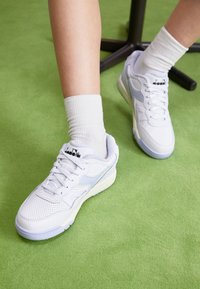 White sneakers with a mesh upper, light blue accents, and textured details. Paired with white ribbed socks on green surface.