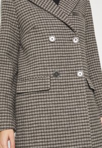 Abrigo de doble botonadura en un patrón de espiga gris y marrón. Fabricado en tela texturizada, con grandes botones blancos y bolsillos laterales.