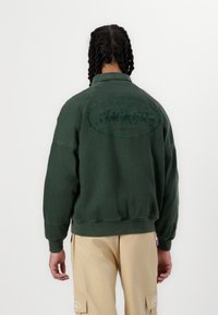 Grøn bomulds sweatshirt med et rundt broderet logo på ryggen. Har ribbede manchetter og kant. Bæres med beige cargo bukser.