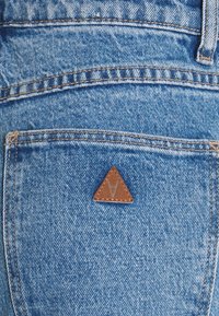 Vaaleansininen farkkukangas, jossa on ruskea kolmionmuotoinen nahkatarra, jossa on logo. Erottuva ommel taskuissa ja saumoissa.