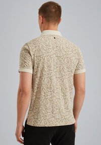 Polo a maniche corte di colore beige con un motivo astratto marrone testurizzato. Presenta un colletto e un dettaglio con bottone sul retro.