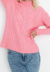 Pull en maille rose avec un col haut et un ourlet côtelé. Porté par-dessus un jean en denim bleu, mettant en valeur une coupe décontractée et un motif texturé.