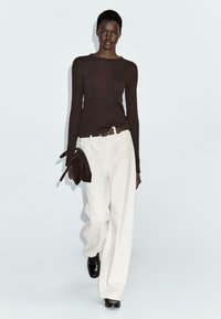Top a maniche lunghe marrone abbinato a pantaloni bianchi a gamba larga. La modella porta una pochette marrone scuro. Tessuto fluido fino al pavimento, design minimalista.
