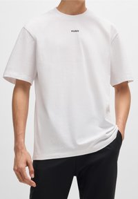 Biała bawełniana koszulka z okrągłym dekoltem i krótkimi rękawami, z małym czarnym logo "HUGO" umieszczonym na środku klatki piersiowej.
