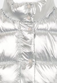 Jachetă puffer metalizată argintie, cu guler înalt, care prezintă butoane cu capsă și o textură matlasată. Suprafață netedă și reflectorizantă, cu un luciu delicat.
