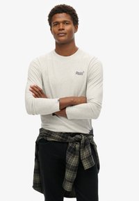 Lichtgrijs langekous t-shirt met een ronde hals en een logo op de borst, gecombineerd met een donkergeruite overhemd dat om de taille is geknoopt.