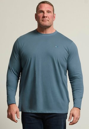 Man with short hair wearing blue long-sleeve shirt and dark pants, standing against plain light background, facing forward.