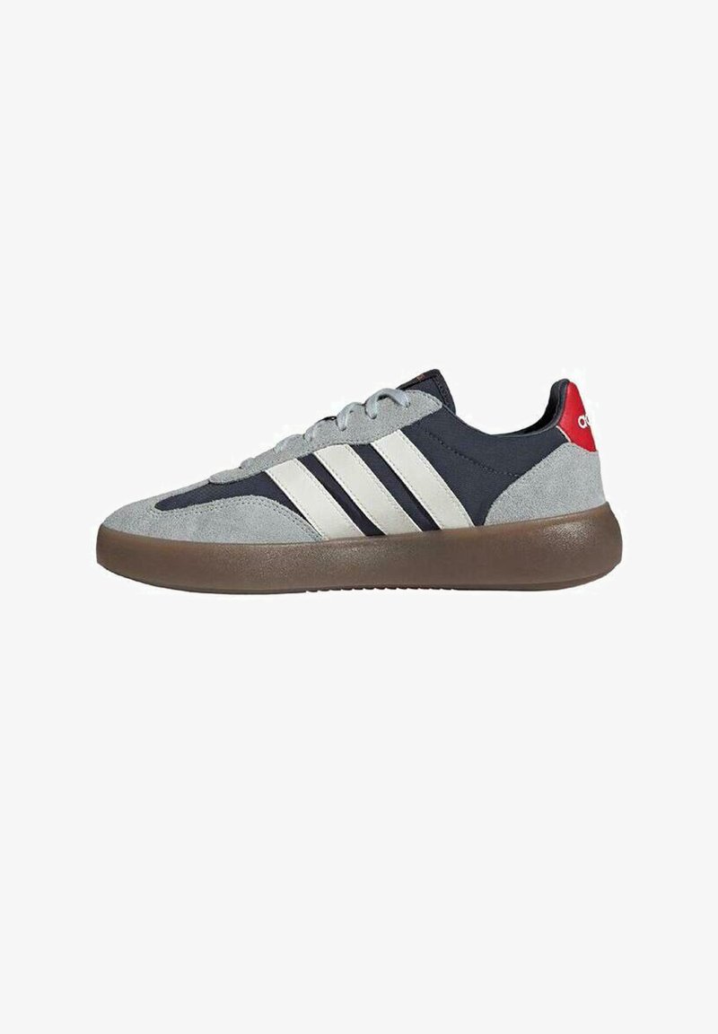 Zapatilla deportiva baja con parte superior de ante gris y tejido azul marino, tres franjas blancas, suela marrón y lengüeta trasera roja con logo.