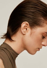 Cabello corto y castaño peinado hacia atrás, primer plano en perfil de un modelo que lleva una blusa marrón de cuello alto; un pequeño arete de perno visible en la oreja.