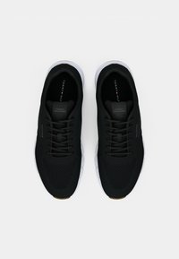 Černé sportovní tenisky s texturovaným síťovaným vrškem, černými šněrovadly, bílou podešví a štítkem s logem na jazyku. Mají zaoblenou špičku.