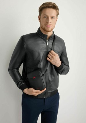 Mężczyzna w czarnej skórzanej kurtce i granatowych spodniach trzyma małą czarną torebkę na pasku na tle jednolitego jasnego tła.