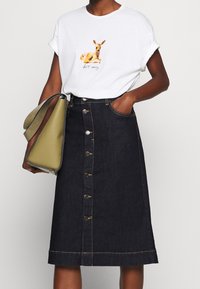 Bílý tričko s grafikou jelena, doplněné tmavými džíny sukní s předními knoflíky a zlatým prošíváním. Zelená kabelka s texturovaným povrchem.