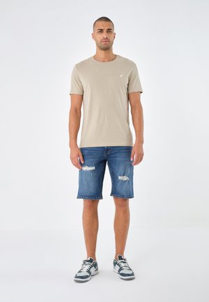 Beige Baumwoll-T-Shirt, blaue ripped Denim-Shorts und graue Sneakers mit weißen Akzenten. Das Modell steht vor einem schlichten Hintergrund.