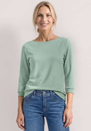 Lächelnde Frau, die ein hellgrünes langärmeliges Oberteil und blaue Jeans trägt, vor einem einfachen hellen Hintergrund stehend.