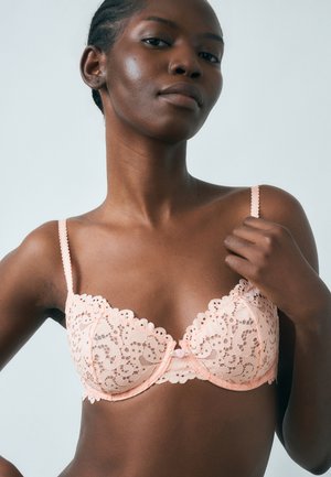 Žena s tmavou pokožkou pózujúca v svetloružovej čipkovanej podprsenke, drží jeden ramienok s neutrálnym výrazom tváre na jednoduchom pozadí.