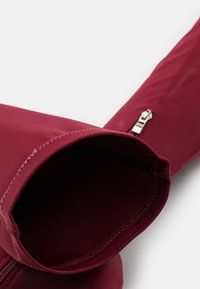 Borsa impermeabile color bordeaux con texture liscia, caratterizzata da una cerniera laterale e cuciture ordinate lungo i bordi, aperta nella parte superiore per un facile accesso.
