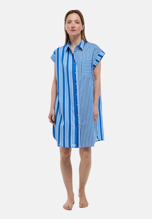 Femme portant une robe chemise rayée verticale bleue et blanche longueur genoux, à manches courtes, debout pieds nus sur un fond uni.