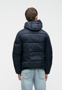 La chaqueta de plumas azul marino cuenta con capucha, diseño acolchado y un grueso aislamiento para mantener el calor. Se lleva con jeans de color azul claro.