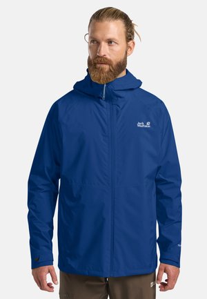 Bärtiger Mann trägt eine blaue wasserdichte Jack Wolfskin Jacke mit Kapuze, steht vor einem einfarbigen Hintergrund.
