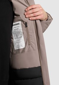 Vékony párnázatú dzseki világosbarna külső résszel és fekete alsó szekcióval, cipzáras zsebbel és belső ápolási címkével.