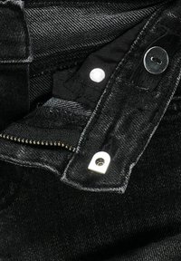 Primo piano di jeans in denim nero che mostrano dettaglio del bottone in metallo, della cerniera e delle cuciture del tessuto.