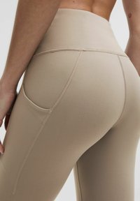 Leggings aderenti color beige a vita alta, con una superficie liscia, dotati di una tasca laterale e cuciture a contrasto lungo le giunture.