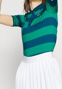 Striped polo shirt in green and navy with a collar and three buttons, paired with a white pleated skirt. Smooth fabric and fitted design.