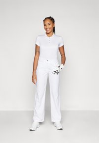 Tricou polo de golf alb cu trei butoane și guler roz deschis, asortat cu pantaloni albi și o mănușă de golf albă, purtat cu pantofi albi.