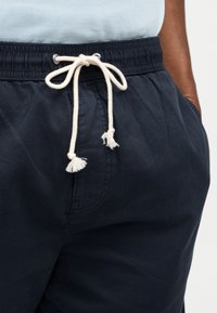 Κοντινό πλάνο σκούρων μπλε παντελονιών με ελαστική μέση και λευκό κορδόνι δεμένο σε φιόγκο, φορεμένα με ανοιχτό μπλε πουκάμισο.