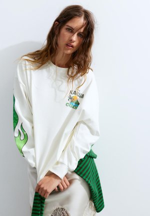 Jeune femme aux longs cheveux bruns porte un sweat-shirt blanc avec des flammes vertes sur les manches et une chemise rayée verte nouée autour de la taille.