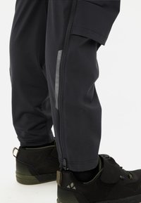 Zwarte sportbroek met een ritssluiting aan de onderkant en een reflecterende strook, gecombineerd met zwarte en groene sneakers met textuurdetails.