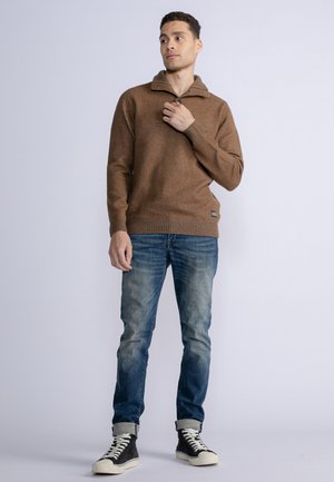 Un pull en laine marron avec un col zippé mi-hauteur, porté avec un jean bleu délavé et des baskets montantes noires. Tissu texturé, coupe décontractée.