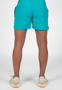 Pantaloni da bagno turchesi con vita elastica e tasche laterali, caratterizzati da una texture liscia e un taglio semplice, indossati con sneaker di colore neutro.