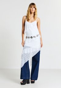 Donna che indossa un top asimmetrico in pizzo bianco con cintura argentata, jeans a gamba larga blu e décolleté neri su uno sfondo neutro.