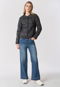 Giacca imbottita trapuntata nera con scollo rotondo e chiusura con bottoni, abbinata a jeans a gamba larga azzurri chiaro. Scarpe da ginnastica bianche completano l'outfit.