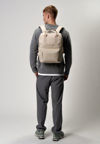 Sac à dos beige avec des bretelles rembourrées, une poignée sur le dessus, une poche zippée à l'avant et une finition texturée. Porté par une personne en pull gris et pantalon gris.