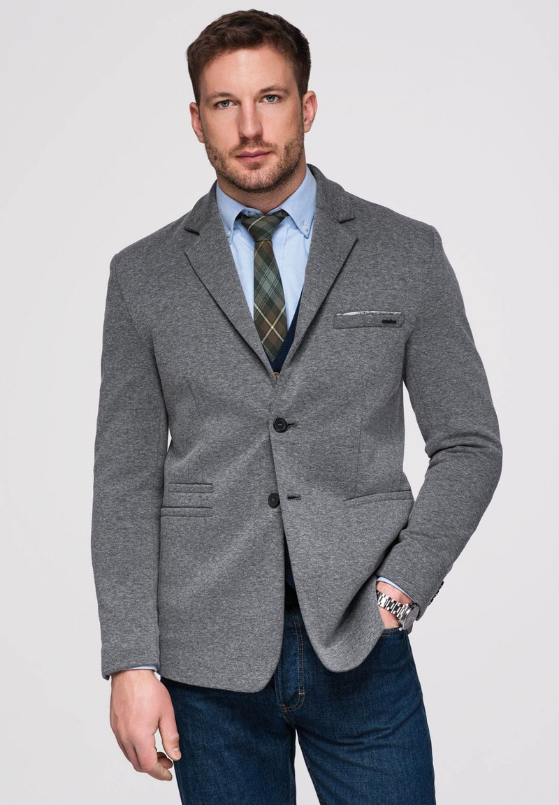 Homme portant un blazer gris, une chemise bleue, une cravate à carreaux, un jean et une montre, debout une main dans la poche sur un fond uni.