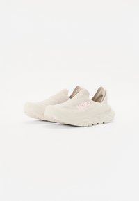 Světlé béžové slip-on tenisky s texturovaným pleteným svrškem, polstrovaným límcem a růžovým logem. Mají flexibilní, tvarovanou podrážku s protiskluzovým vzorem.