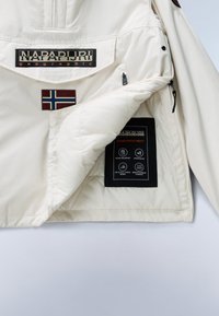 Weiße Napapijri-Jacke mit vorderer Tasche mit Logo, norwegischem Flaggen-Patch und Innenetikett, das Wasserbeständigkeit und atmungsaktiven Stoff hervorhebt.