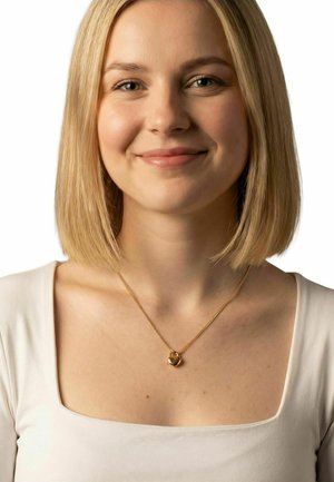 Jonge vrouw met blond bobkapsel, draagt een wit topje met een vierkante halslijn en een gouden hartvormige hangers ketting, glimlacht zachtjes.